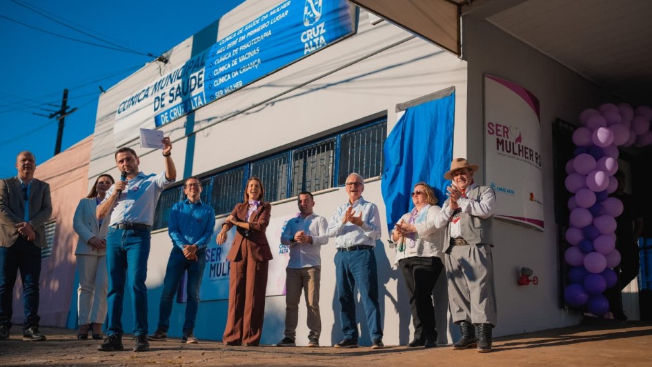 O vice-governador Gabriel Souza aparece em primeiro plano, de camiseta branca e jeans, discursando ao microfone durante a inauguração do 13º Serviço Especializado de Referência à Saúde da Mulher (SERMulher RS), em Cruz Alta. Ao lado dele, em frente ao prédio identificado como Clínica Municipal de Saúde de Cruz Alta, estão autoridades estaduais e municipais, homens e mulheres, que aplaudem e sorriem. À direita da imagem, próximo à entrada do prédio, há uma faixa do programa Ser Mulher RS e uma decoração com balões lilás. O evento ocorre em área externa, sob céu azul.