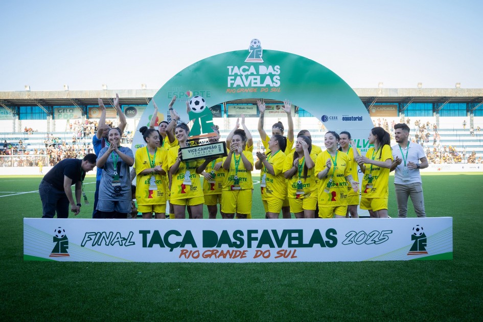 A imagem mostra um grupo de jogadoras vestidas com uniformes esportivos amarelos comemorando em um campo de futebol. Elas estão segurando um troféu de vice-campeãs e posicionadas atrás de uma faixa com os dizeres "Final Taça das Favelas 2025 Rio Grande do Sul". Ao fundo, há espectadores nas arquibancadas.