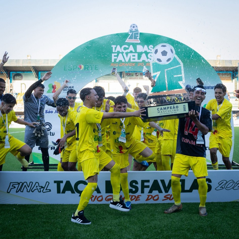 A imagem mostra um grupo de jogadores de futebol comemorando a vitória na Taça das Favelas, vestindo uniformes amarelos e reunidos em torno de um grande troféu com a inscrição "Campeão Masculino 2025". Eles estão posicionados diante de um arco verde com os dizeres "Taça das Favelas" e logotipos de patrocinadores.