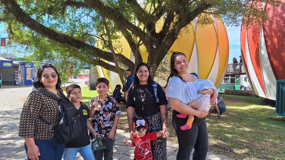 Três mulheres e quatro crianças posam para a foto embaixo de uma árvore, no Parque Assis Brasil. Ao fundo, aparecem as tradicionais esferas nas cores amarela e vermelha da Expointer.