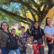 Três mulheres e quatro crianças posam para a foto embaixo de uma árvore, no Parque Assis Brasil. Ao fundo, aparecem as tradicionais esferas nas cores amarela e vermelha da Expointer. 