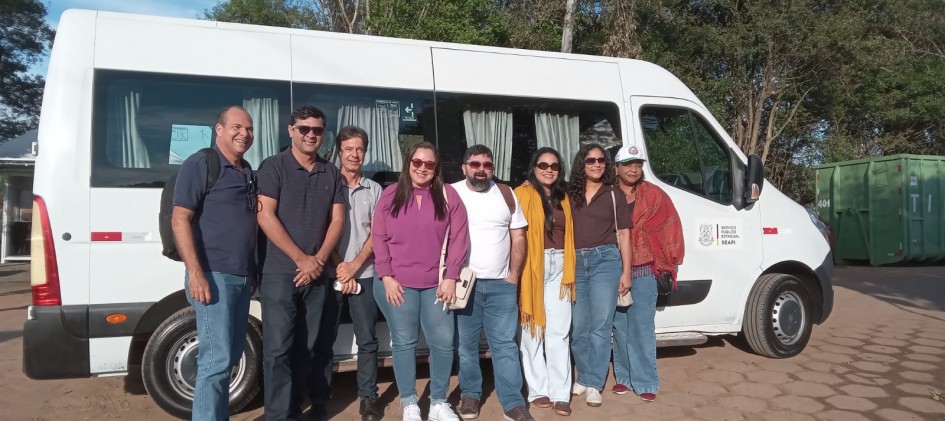 Oito pessoas posam lado a lado, em pé, sorrindo para a câmera. Elas estão no sol, posicionadas em frente a uma van.
