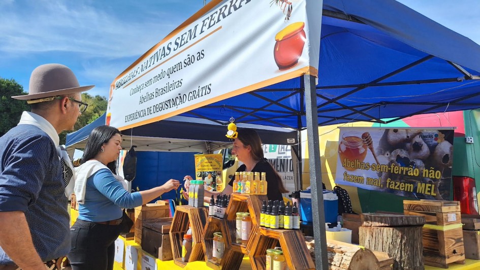 Um homem e uma mulher estão posicionados ao lado esquerdo da imagem. Eles estão em frente a um estande com produtos de abelhas nativas sem ferrão. Há diversos itens, como mel, própolis e cera.