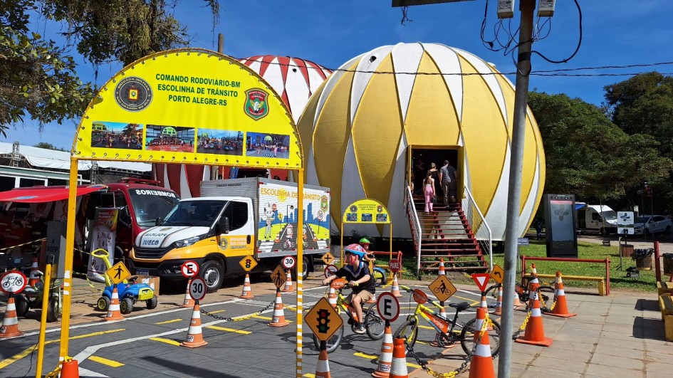 Em um ambiente ao ar livre, há uma mini pista montada com cones e placas de sinalização de trânsito. Há uma criança andando de bicicleta no trecho. Ao fundo aparecem grandes esferas de cor amarela e vermelha da Expointer e um caminhão identificando a Escolinha de Trânsito do Comando Rodoviário da Brigada Militar.