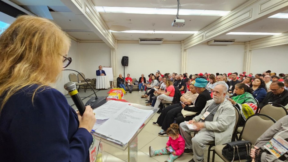 Mulher loira, de óculos, fala ao microfone em um auditório lotado. Ela está de costas para a câmera, lendo de uma folha sobre um púlpito transparente. À frente, diversas pessoas estão sentadas, entre elas adultos, idosos e uma criança sentada no chão. No palco, um homem está sentado à mesa, observando a apresentação. O ambiente é iluminado e possui teto branco com projetor instalado.