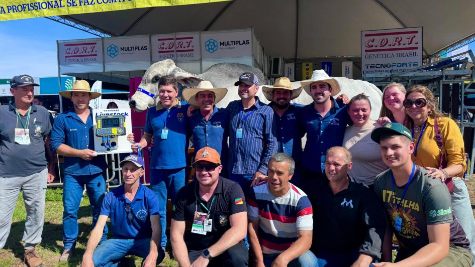 Na imagem, um grupo de aproximadamente 15 pessoas posa para foto em frente a um estande na Expointer. Entre os participantes, estão homens e mulheres, alguns usando chapéus de palha ou bonés, e muitos com crachás de identificação pendurados no pescoço. Ao fundo, é possível ver um grande boi branco da raça zebuína, identificado por uma faixa azul na cabeça, em destaque como atração principal. Um dos homens segura um painel eletrônico que marca “1430”, indicando o peso do animal, que simboliza o recorde de maior bovino já apresentado na feira. O ambiente é de celebração e registro histórico.