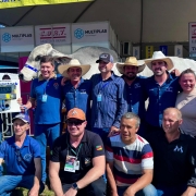 Na imagem, um grupo de aproximadamente 15 pessoas posa para foto em frente a um estande na Expointer. Entre os participantes, estão homens e mulheres, alguns usando chapéus de palha ou bonés, e muitos com crachás de identificação pendurados no pescoço. Ao fundo, é possível ver um grande boi branco da raça zebuína, identificado por uma faixa azul na cabeça, em destaque como atração principal. Um dos homens segura um painel eletrônico que marca “1430”, indicando o peso do animal, que simboliza o recorde de maior bovino já apresentado na feira. O ambiente é de celebração e registro histórico.