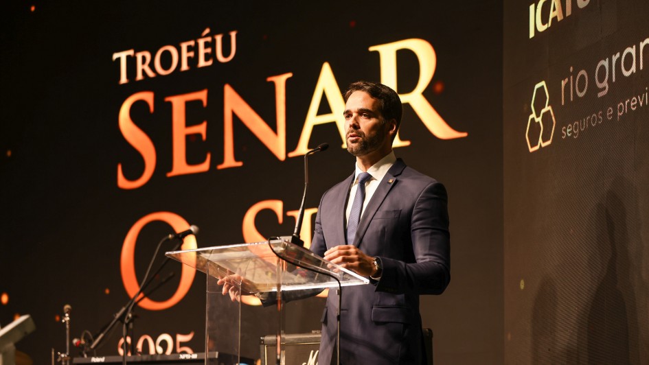 Foto em plano americano de Leite discursando no púlpito. Ao fundo, um grande painel preto com os dizeres em dourado: Troféu Senar o Sul.