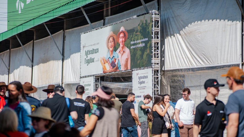 A imagem mostra um evento ao ar livre com várias pessoas caminhando e interagindo em frente a um grande estande. Na parte superior do estande há uma placa verde com o logotipo do Sicredi. Abaixo, há um banner grande com a foto de duas pessoas sorridentes (um homem e uma mulher mais velhos, com roupas típicas de agricultores), segurando cestas com produtos agrícolas como pão, uvas e verduras. No banner está escrito:
"29ª Expointer 2025
Agricultura Familiar
Mais do que produtos: amor e tradição".
Muitas pessoas estão passando ou paradas em frente ao estande. A maioria veste roupas casuais de verão e alguns usam chapéus para se proteger do sol. O ambiente parece ser uma feira ou exposição agropecuária, com clima descontraído e ensolarado.