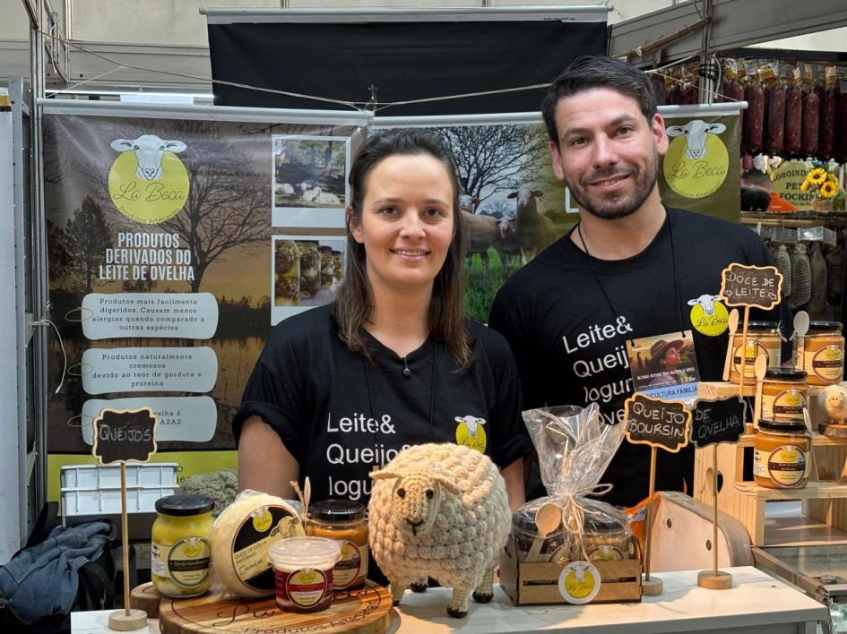 Fotografia de dois expositores em uma feira de produtos artesanais, posicionados atrás de um balcão decorado com itens derivados de leite de ovelha. À esquerda, uma mulher de cabelos castanhos presos sorri para a câmera; à direita, um homem de barba curta também sorri. Ambos vestem camisetas pretas com os dizeres em branco: “Leite& Queijo& Iogurte&”.
Sobre a bancada estão dispostos potes de queijos, doce de leite, iogurtes e um queijo embalado a vácuo. Há também uma pequena ovelha de pelúcia bege como elemento decorativo. Placas de madeira indicam os produtos, como “Queijos”, “Doce de Leite” e “Queijo Boursin de Ovelha”.
Ao fundo, um banner da marca “La Beca – Produtos Artesanais” traz a imagem de uma ovelha e os dizeres “Produtos derivados do leite de ovelha”. O ambiente é de feira, com outros estandes e produtos visíveis ao redor.