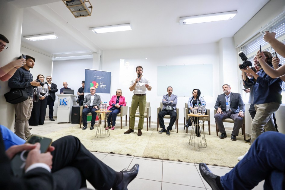 A imagem mostra um evento oficial sendo realizado em um ambiente fechado, com diversas pessoas presentes.
No centro da cena, um homem de barba, vestindo camisa branca e calça verde, está em pé, segurando um microfone e falando ao público. Atrás dele, há cinco pessoas sentadas em cadeiras, compondo uma mesa de autoridades. Entre elas, uma mulher de blazer rosa-choque (à esquerda do orador), e outras pessoas com trajes formais.
À esquerda, há uma bancada com o logotipo do Hospital Santa Cruz (HSC) e um banner da Secretaria da Saúde do Estado do Rio Grande do Sul, indicando que o evento está relacionado à saúde pública. Várias pessoas assistem ao evento de pé, inclusive jornalistas com câmeras e celulares à direita da imagem, registrando o momento.
O ambiente tem paredes brancas, iluminação fluorescente e janelas com persianas. O chão é revestido com tapete claro na área da frente.