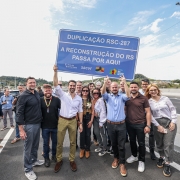 A imagem mostra um grupo de pessoas reunidas sobre uma rodovia. No centro, duas pessoas seguram uma placa azul com os dizeres: "Duplicação RSC-287 - A reconstrução do RS passa por aqui". 