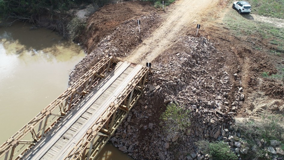 A imagem mostra uma ponte de madeira estreita sobre um rio de água barrenta. As cabeceiras da ponte foram reforçadas com pedras e terra, formando rampas de acesso. No lado direito, há uma estrada de chão batido onde estão dois carros brancos estacionados e algumas pessoas em pé, observando a obra. Ao fundo, há vegetação densa.