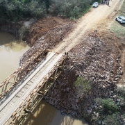 A imagem mostra uma ponte de madeira estreita sobre um rio de água barrenta. As cabeceiras da ponte foram reforçadas com pedras e terra, formando rampas de acesso. No lado direito, há uma estrada de chão batido onde estão dois carros brancos estacionados e algumas pessoas em pé, observando a obra. Ao fundo, há vegetação densa.
