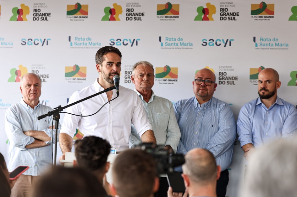 A imagem mostra cinco pessoas em pé diante de um painel com logotipos e textos institucionais, incluindo "Governo do Estado do Rio Grande do Sul", "Plano Rio Grande", "Rota de Santa Maria" e "Sacyr". O governador está falando ao microfone em um púlpito, enquanto as demais acompanham ao fundo.