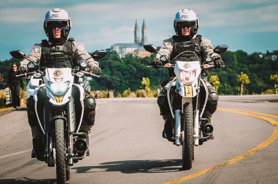 Foto mostra dois policiais militares andando em duas motos, numa curva de uma estrada. Eles estão com capacetes brancos, uniformes beges e coletes pretos e joelheiras pretas. Ao fundo, há uma mata verde e bem ao fundo a silhueta de uma igreja com duas torres pontiaguadas, no alto da colina.