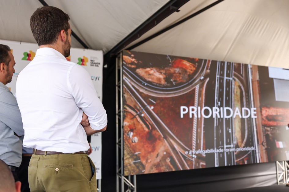 Duas pessoas observam uma tela grande sob uma tenda. A imagem mostra um cruzamento de rodovias visto de cima, com a palavra “PRIORIDADE” em destaque. É um evento sobre infraestrutura e mobilidade urbana.