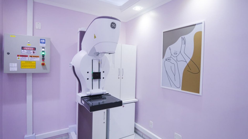 Descrição para fins de acessibilidade: Sala de exames com paredes lilás, piso de cerâmica clara e iluminação embutida no teto. No centro, há um equipamento de mamografia de cor branca e detalhes em roxo, com a logomarca da GE na parte superior. À esquerda, vê-se um painel elétrico cinza com avisos de “Perigo” em vermelho e amarelo. Ao fundo, há um armário branco. Na parede à direita, está pendurado um quadro com ilustração minimalista em traços pretos de uma mulher em perfil, com fundo em tons bege e cinza.