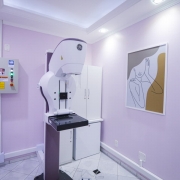 Descrição para fins de acessibilidade: Sala de exames com paredes lilás, piso de cerâmica clara e iluminação embutida no teto. No centro, há um equipamento de mamografia de cor branca e detalhes em roxo, com a logomarca da GE na parte superior. À esquerda, vê-se um painel elétrico cinza com avisos de “Perigo” em vermelho e amarelo. Ao fundo, há um armário branco. Na parede à direita, está pendurado um quadro com ilustração minimalista em traços pretos de uma mulher em perfil, com fundo em tons bege e cinza.