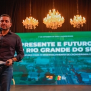 Um homem está em pé sobre um palco, segurando um microfone com uma mão e uma folha de papel com a outra. Ele veste uma camisa preta e jeans. Ao fundo, há um grande telão com os dizeres: "Presente e Futuro do Rio Grande do Sul – Estratégias para o desenvolvimento de Cachoeirinha e região". No topo do telão, está a data do evento: "1º de setembro de 2023 | Cachoeirinha". Acima da tela, três lustres de cristal iluminam o ambiente, sugerindo que o evento ocorre em um teatro ou auditório. O clima da imagem é formal, com foco em uma apresentação institucional.