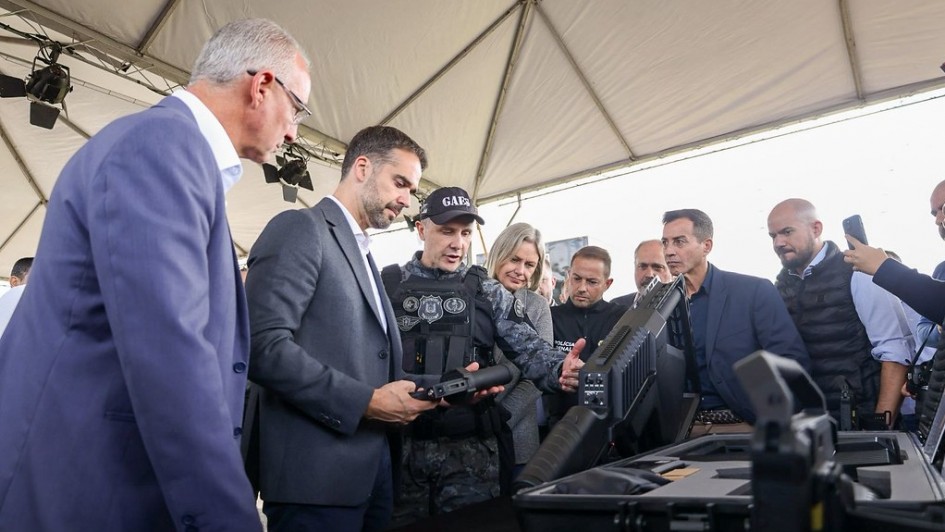 Foto mostra o governador Eduardo Leite em um estande onde são expostos armamentos novos para a Policia Penal. Ele está olhando para um armamento que está em sua mão. Ao seu lado, um policial penal, fardado, com um boné preto e coletes pretos, explica sobre outra arma, um fuzil, que está sobre uma mesa. Ao seu redor, outras autoridades acompanham a cena atentamente.