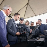 Foto mostra o governador Eduardo Leite em um estande onde são expostos armamentos novos para a Policia Penal. Ele está olhando para um armamento que está em sua mão. Ao seu lado, um policial penal, fardado, com um boné preto e coletes pretos, explica sobre outra arma, um fuzil, que está sobre uma mesa. Ao seu redor, outras autoridades acompanham a cena atentamente. 