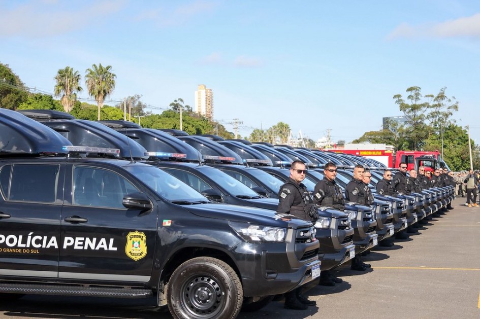 Foto mostra uma frota de viaturas da Polícia Penal, com policiais perfilados à frente delas. As viaturas são pretas e os policiais vestem uniformes pretos e usam óculos escuros pretos. Na viatura mais próxima, aparece o nome da Polícia Penal e o distintivo, de cor amarela.
