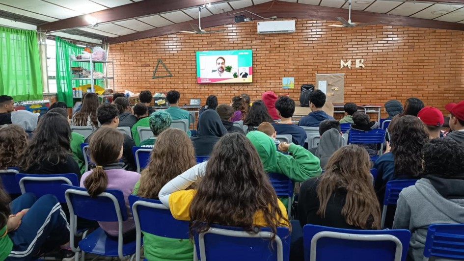 Em uma sala, diversos estudantes estão reunidos, sentados em cadeiras, para assistir a live projetada em uma TV no centro da imagem. Os estudantes estão de costas e na tela aparece o governador Eduardo Leite falando.