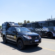 Foto mostra uma viatura da Polícia Penal em primeiro plano, na cor preta, com emblema em amarelo. Ao fundo, há um ônibus, com estampa camuflada azul e cinza, com a palavra "choque" em destaque na cor branca.  