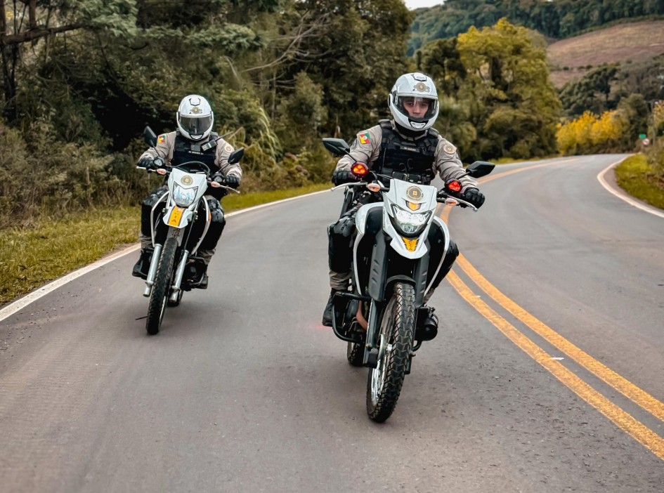 Foto mostra dois policiais militares em duas motos, com frente branca. Eles estão numa curva de uma estrada de asfalto. Os dois estão de uniforme bege, colete preto e capacete branco.