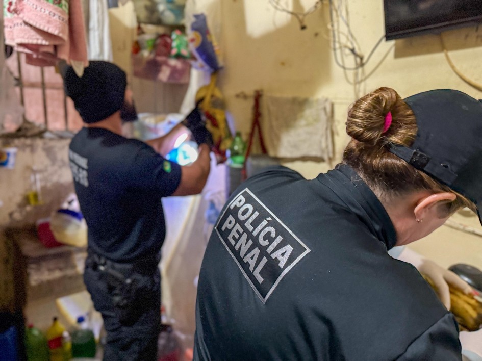 Foto mostra dois policiais penais, com uniformes preto, que estão de costas, fazendo uma vistoria dentro de uma cela, de paredes amarelas. O local é cheio de objetos que estão desfocados.