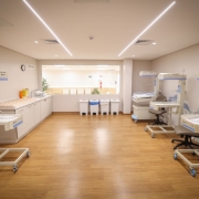 Descrição para fins de acessibilidade: Sala do centro obstétrico de um hospital, ampla, com piso amadeirado e iluminação em linhas de LED no teto. À direita, três estações de cuidado neonatal com berços aquecidos transparentes sobre rodízios e cadeiras giratórias. À esquerda, outra estação semelhante ao lado de uma bancada com pia, dispensers e um relógio de parede. No fundo, há uma janela interna para o corredor e cinco lixeiras com tampas coloridas para separação de resíduos. O ambiente está vazio, organizado e pronto para uso.