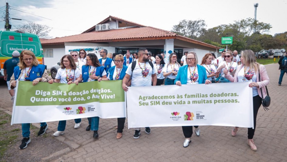 Foto em ambiente aberto. Um grupo de pessoas de camisetas brancas e coletes azuis caminha segurando cartazes relacionados à campanha para doação de órgãos.