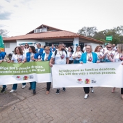 Foto em ambiente aberto. Um grupo de pessoas de camisetas brancas e coletes azuis caminha segurando cartazes relacionados à campanha para doação de órgãos. 