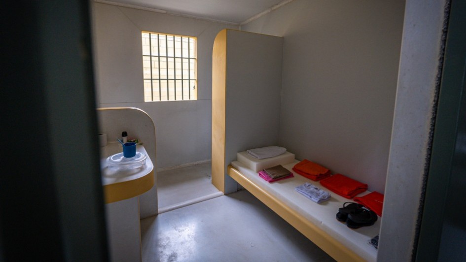 Cela de uma prisão, mostrando uma cama, com as roupa de cama branca e sobre ela itens como roupas para os detentos, na cor laranja. A cela tem piso cinza e paredes brancas com detalhes em amarelo. Ao fundo, uma janela com grades.