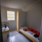 Cela de uma prisão, mostrando uma cama, com as roupa de cama branca e sobre ela itens como roupas para os detentos, na cor laranja. A cela tem piso cinza e paredes brancas com detalhes em amarelo. Ao fundo, uma janela com grades.
