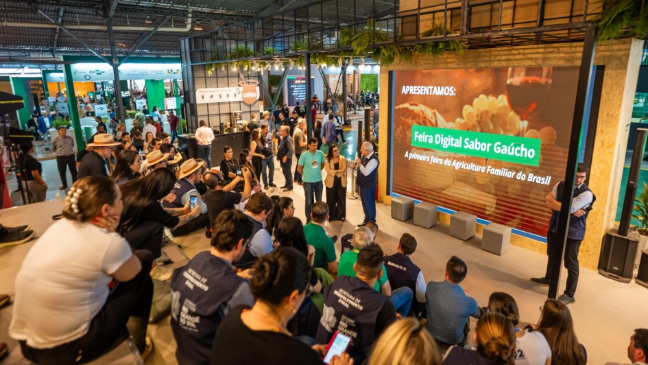 A foto mostra um evento em ambiente interno, possivelmente uma feira ou apresentação. Um grupo de pessoas, algumas sentadas no chão e outras em um banco de madeira em primeiro plano, está assistindo a uma apresentação. A maioria das pessoas veste camisas com o logo "Feira Digital Sabor Gaúcho" nas costas, e algumas usam chapéus de palha. Em frente a eles, três pessoas, incluindo um homem de blazer azul e calça cinza, estão em pé conversando. Um grande telão ao fundo mostra uma imagem de queijo e vinho, com o texto "APRESENTAMOS: Feira Digital Sabor Gaúcho, a primeira feira da Agricultura Familiar do Brasil". A iluminação é mista, com luzes de LED no teto de estrutura metálica.