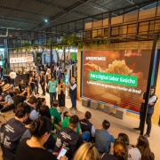 A foto mostra um evento em ambiente interno, possivelmente uma feira ou apresentação. Um grupo de pessoas, algumas sentadas no chão e outras em um banco de madeira em primeiro plano, está assistindo a uma apresentação. A maioria das pessoas veste camisas com o logo "Feira Digital Sabor Gaúcho" nas costas, e algumas usam chapéus de palha. Em frente a eles, três pessoas, incluindo um homem de blazer azul e calça cinza, estão em pé conversando. Um grande telão ao fundo mostra uma imagem de queijo e vinho, com o texto "APRESENTAMOS: Feira Digital Sabor Gaúcho, a primeira feira da Agricultura Familiar do Brasil". A iluminação é mista, com luzes de LED no teto de estrutura metálica.