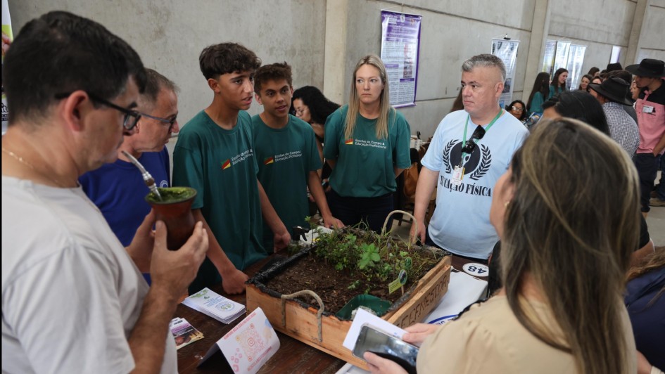Na imagem, um grupo de estudantes e professores está reunido em torno de uma mesa de exposição em um espaço coberto. Sobre a mesa há uma caixa de madeira com plantas cultivadas em terra, representando um projeto escolar ligado à agricultura ou sustentabilidade. Dois jovens com camisetas verdes da Escola do Campo apresentam a atividade, enquanto adultos escutam atentos. Entre eles, um homem segura uma cuia de chimarrão e outro usa camiseta azul clara escrita “Educação Física”. A cena transmite aprendizado coletivo e valorização do conhecimento local.