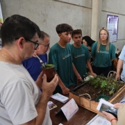Na imagem, um grupo de estudantes e professores está reunido em torno de uma mesa de exposição em um espaço coberto. Sobre a mesa há uma caixa de madeira com plantas cultivadas em terra, representando um projeto escolar ligado à agricultura ou sustentabilidade. Dois jovens com camisetas verdes da Escola do Campo apresentam a atividade, enquanto adultos escutam atentos. Entre eles, um homem segura uma cuia de chimarrão e outro usa camiseta azul clara escrita “Educação Física”. A cena transmite aprendizado coletivo e valorização do conhecimento local.