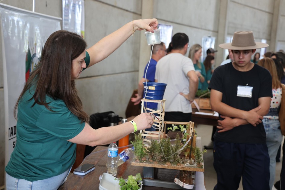 A foto mostra uma mulher com camiseta verde demonstrando um experimento em uma feira escolar. Ela segura um copo transparente com água, conectado por uma mangueira a um pequeno sistema de irrigação feito com balde azul e suporte de madeira. Sobre a mesa, plantas pequenas estão dispostas em um modelo que simula técnicas agrícolas sustentáveis. Um jovem de chapéu observa atentamente a explicação, enquanto outras pessoas circulam pelo ambiente. A cena destaca práticas educativas de ciência aplicada à vida rural.