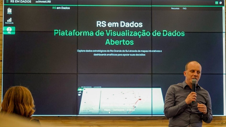 A imagem mostra uma apresenta&ccedil;&atilde;o em um ambiente interno, provavelmente um audit&oacute;rio ou sala de confer&ecirc;ncias. No centro da imagem, h&aacute; uma grande tela dividida em v&aacute;rios pain&eacute;is exibindo a plataforma "RS em Dados", com o texto:

"RS em Dados &ndash; Plataforma de Visualiza&ccedil;&atilde;o de Dados Abertos. Explore dados estrat&eacute;gicos do Rio Grande do Sul atrav&eacute;s de mapas interativos e dashboards anal&iacute;ticos para apoiar suas decis&otilde;es."

&Agrave; direita da imagem, est&aacute; um homem careca, vestindo camisa cinza, segurando um microfone enquanto fala. Ele parece estar apresentando ou explicando a plataforma. &Agrave; esquerda, de costas, est&aacute; uma mulher assistindo &agrave; apresenta&ccedil;&atilde;o.

O ambiente sugere uma ocasi&atilde;o formal de lan&ccedil;amento ou demonstra&ccedil;&atilde;o de um sistema digital governamental. A interface da plataforma exibida na tela tem fundo escuro com textos e elementos gr&aacute;ficos em verde e branco.