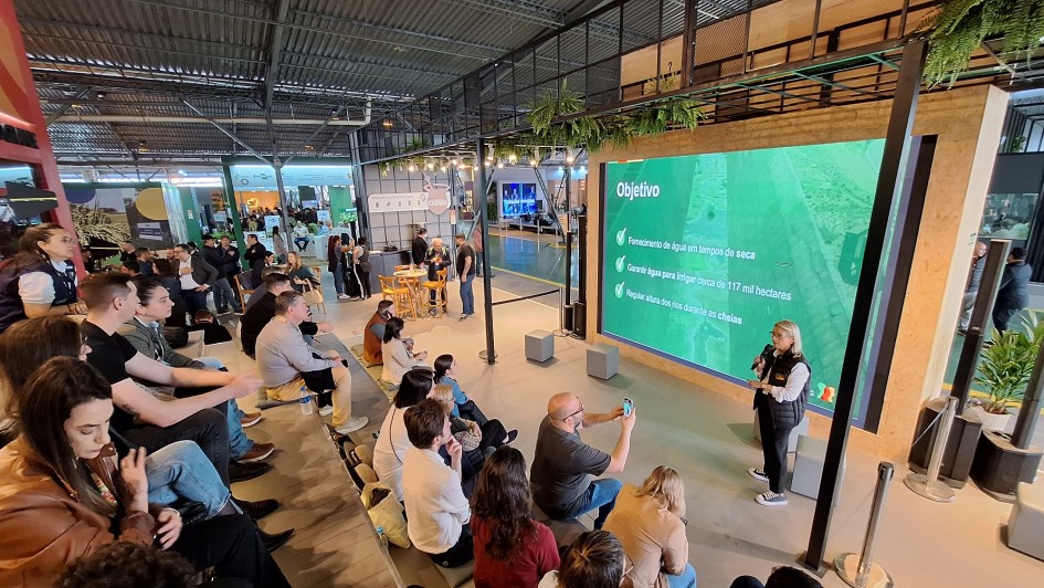 Imagem mostra uma palestra realizada em espa&ccedil;o fechado, com arquibancada ocupada por diversas pessoas que acompanham a apresenta&ccedil;&atilde;o. No palco, uma mulher segura microfone e fala diante de um grande tel&atilde;o verde que exibe o t&iacute;tulo &ldquo;Objetivo&rdquo; e tr&ecirc;s t&oacute;picos: fornecimento de &aacute;gua em tempos de seca; garantia de irriga&ccedil;&atilde;o para cerca de 117 mil hectares; e regula&ccedil;&atilde;o da altura dos rios durante as cheias. O ambiente &eacute; moderno, com estrutura met&aacute;lica, ilumina&ccedil;&atilde;o artificial e estandes ao fundo.