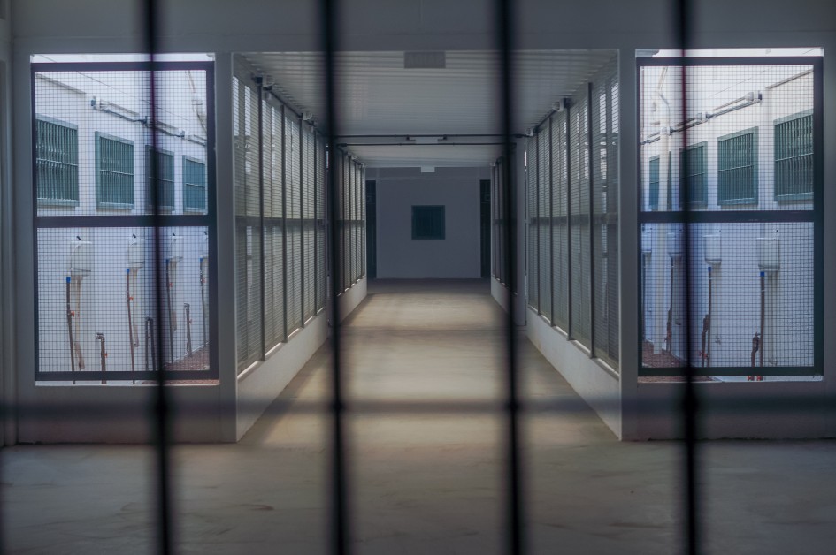 A foto mostra, por dentro dos vãos de uma grade, um corredor de uma galeria de uma unidade prisional.