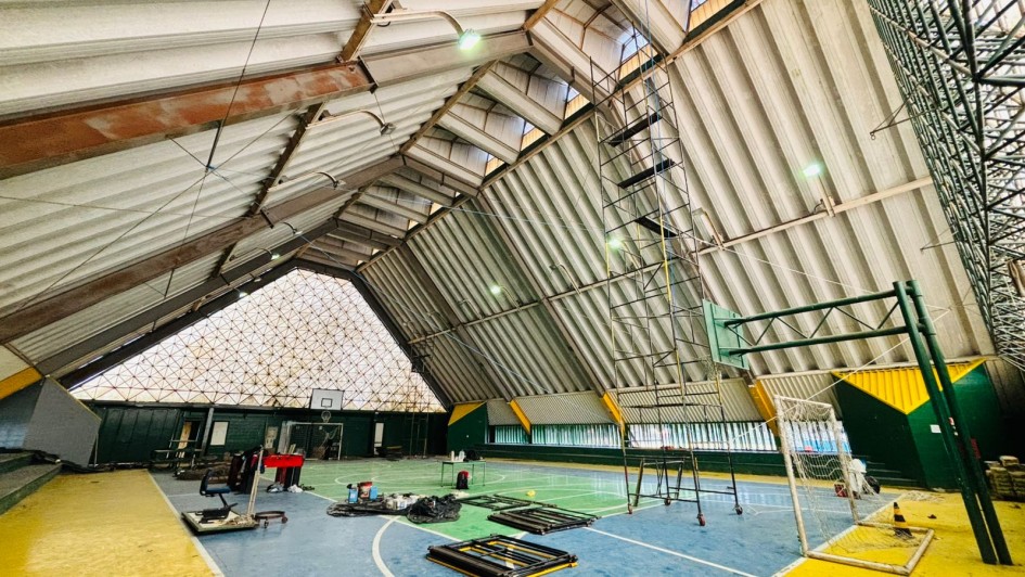 Foto interna de um ginásio, com teto triangular alto. A quadra está pintada de verde, azul e amarelo. Há andaimes e ferramentas que indicam obra em andamento na estrutura.