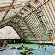Foto interna de um ginásio, com teto triangular alto. A quadra está pintada de verde, azul e amarelo. Há andaimes e ferramentas que indicam obra em andamento na estrutura. 