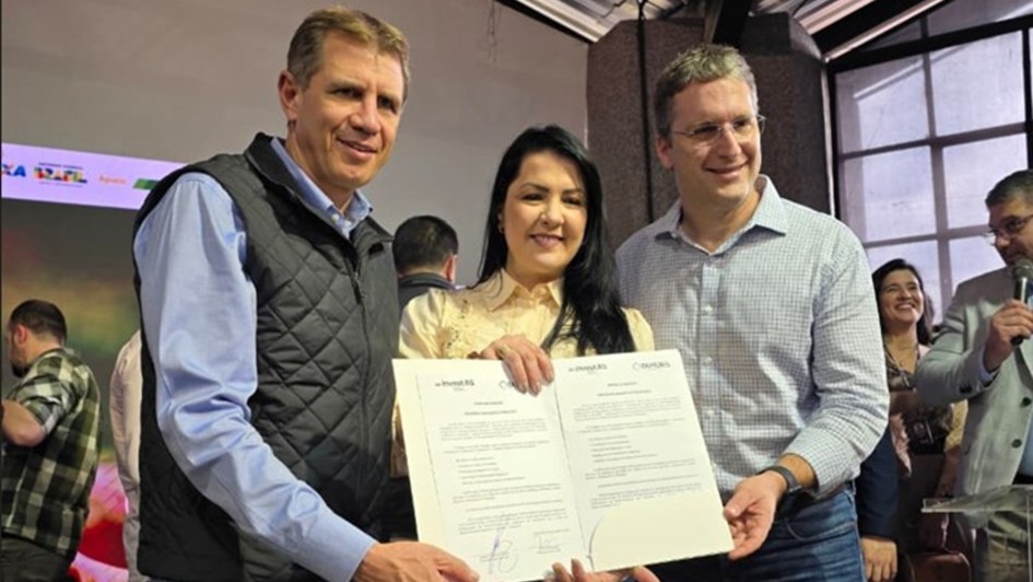 Três pessoas estão em destaque no centro da imagem, sorrindo e segurando um documento oficial assinado. À esquerda, um homem de colete preto e camisa azul clara; ao centro, uma mulher de cabelos longos e escuros, vestindo uma blusa clara; e à direita, outro homem de óculos, com camisa xadrez azul clara. O documento que seguram possui os logotipos do "Invest.RS" e "Taurus" no topo. Ao fundo, há outras pessoas participando do evento, uma delas falando ao microfone. O ambiente parece ser um espaço fechado, possivelmente um auditório ou salão de eventos, com iluminação natural entrando pelas janelas.