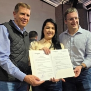 Três pessoas estão em destaque no centro da imagem, sorrindo e segurando um documento oficial assinado. À esquerda, um homem de colete preto e camisa azul clara; ao centro, uma mulher de cabelos longos e escuros, vestindo uma blusa clara; e à direita, outro homem de óculos, com camisa xadrez azul clara. O documento que seguram possui os logotipos do "Invest.RS" e "Taurus" no topo. Ao fundo, há outras pessoas participando do evento, uma delas falando ao microfone. O ambiente parece ser um espaço fechado, possivelmente um auditório ou salão de eventos, com iluminação natural entrando pelas janelas.