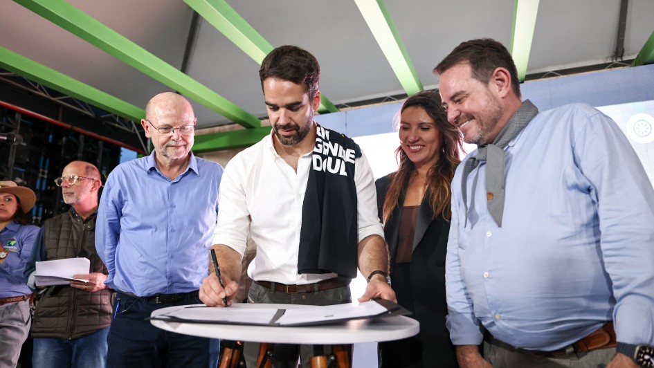 Em um palco coberto, com vigas verdes aparentes, um homem de camisa branca assina um documento sobre uma mesa redonda alta, de tampo branco e pés de madeira. Ao redor dele, quatro pessoas observam sorrindo — dois homens à esquerda e, à direita, uma mulher e outro homem em traje social. Um tecido preto com letras brancas está apoiado no ombro do signatário. Ao fundo, há um telão iluminado e equipamentos de evento.
