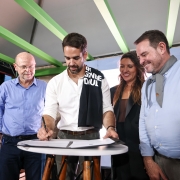 Em um palco coberto, com vigas verdes aparentes, um homem de camisa branca assina um documento sobre uma mesa redonda alta, de tampo branco e pés de madeira. Ao redor dele, quatro pessoas observam sorrindo — dois homens à esquerda e, à direita, uma mulher e outro homem em traje social. Um tecido preto com letras brancas está apoiado no ombro do signatário. Ao fundo, há um telão iluminado e equipamentos de evento.